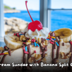 Close-up of an ice cream sundae with banana slices, nuts, chocolate drizzle, whipped cream, sprinkles, and a cherry on top, with a seaside view in the background.