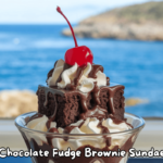 A rich chocolate fudge brownie sundae topped with whipped cream, chocolate drizzle, and a cherry, set against a scenic ocean backdrop.